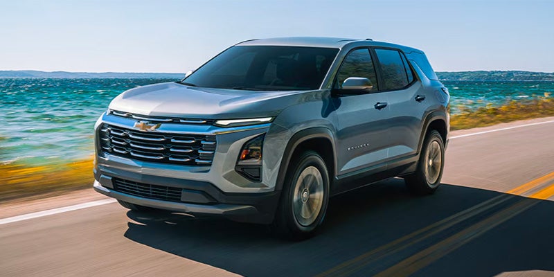 A gray Chevrolet SUV drives on a coastal highway with a blue ocean and distant land visible to the left