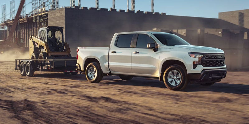White Chevrolet Silverado pickup truck towing a yellow skid-steer loader on a flatbed trailer at a construction site