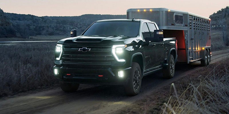 Dark gray pickup truck with headlights on, pulling a horse trailer down a dirt road at dusk