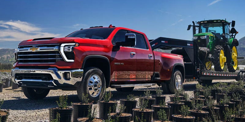 Red Chevrolet 3500 HD truck towing a green John Deere tractor on a trailer, parked among potted plants