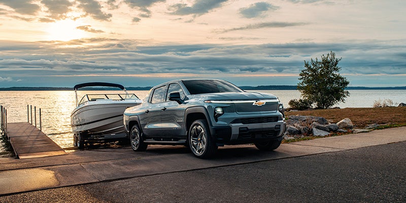 A gray electric Chevy Silverado truck towing a white boat on a trailer, backed into a boat launch at sunset