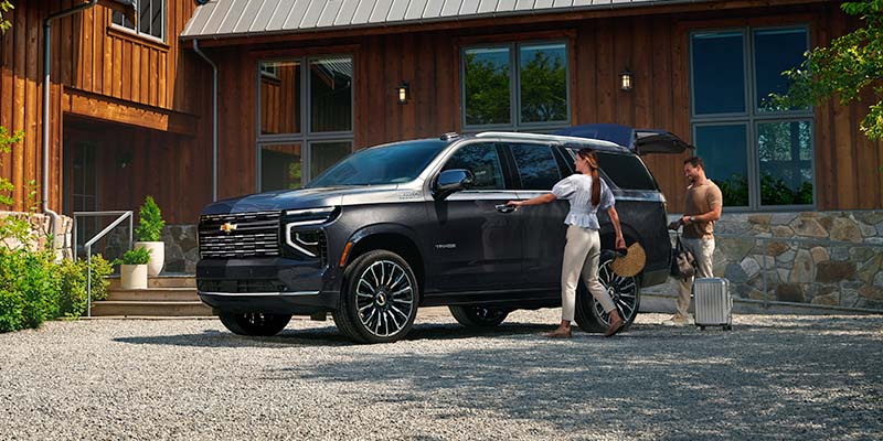 A dark gray Chevrolet Tahoe SUV parked on a gravel driveway with a couple loading luggage