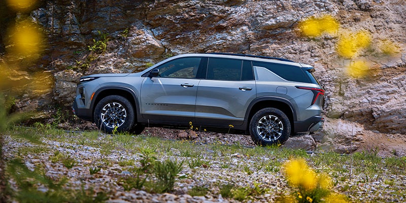 A gray Traverse SUV with black wheels with silver accents is parked on a rocky incline next to a cliff face