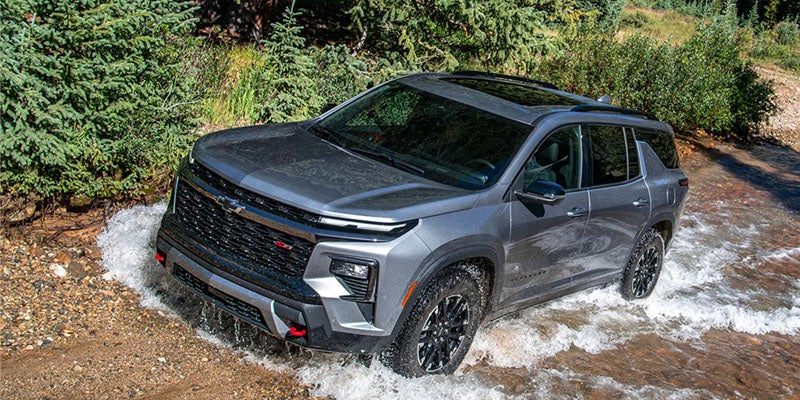 A gray Chevrolet Traverse Z71 SUV driving through a shallow, rocky river with trees in the background